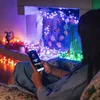 A person is using a smartphone to control a string of colorful LED lights that are decorating a fireplace and a window. Náhled obrázku produktu