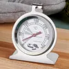 A close-up shot of a stainless steel oven thermometer with a stand, placed on a wooden surface. A grey fabric item is visible in the background. Produktbild-Vorschau