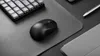 A black wireless mouse is placed on a black mousepad next to a black keyboard and a silver stylus pen. Produktbild-Vorschau