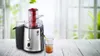 A juicer is shown on a wooden table with apples in the feeder and juice being poured into a glass. The background is a blurred window with greenery outside. Náhled obrázku produktu