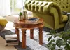 A round wooden coffee table with books, a plant, and a vase on it, next to a green tufted sofa. Produktbild-Vorschau
