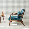A teal velvet armchair with wooden legs is placed against a white wall with a black grid pattern. A small side table with a glass bottle and a bowl is next to the chair. Anteprima dell’immagine prodotto
