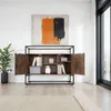 A modern sideboard with industrial-style metal framing and dark wood-effect panels, displayed in a well-lit room with a large window and abstract art on the wall. Náhled obrázku produktu