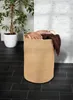 A laundry basket filled with towels is placed on a dark tiled floor next to a potted plant and a white brick wall. Náhled obrázku produktu