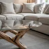 A close-up of a glass-top coffee table with a unique wooden base, placed in a living room setting with a sectional sofa in the background. Produktbild-Vorschau