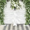 A white fluted pedestal stands against a white brick wall, adorned with lush green foliage and white flowers on either side, creating an elegant archway. A dark urn filled with white flowers sits atop the pedestal. Anteprima dell’immagine prodotto