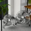 Two metal floor fans are shown on a carpeted floor, with a plant and a shelf in the background. Náhľad obrázku produktu