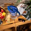 A BRITA water filter pitcher is shown on a wooden table with fruit, a glass of water, a magazine, and sunglasses. Produktbild-Vorschau