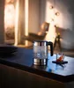 A glass electric kettle is boiling water on a dark countertop, with a small glass of tea next to it. The background is softly lit, suggesting a cozy home environment. Náhled obrázku produktu