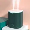 A dark green humidifier with two streams of mist emanating from the top, placed on a white pedestal against a pink and teal background. Produktbild-Vorschau