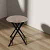 A small round folding stool with a wooden top and black metal legs sits on a wooden floor next to a white wall with shadows cast on it. Produktbild-Vorschau