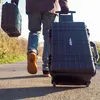 A person walks down a paved path carrying two black cases, one in hand and one with wheels being pulled. Produktbild-Vorschau