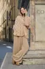 A woman wearing a beige knitted outfit stands on a street in front of a stone structure. Produktbild-Vorschau