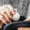 A person is placing a white geo tag on a keychain into a black zippered pouch. Voir les images du produit