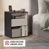 A modern black and gray nightstand with a charging phone, a mug, books, and a storage basket, placed next to a bed. Produktbild-Vorschau