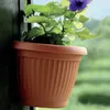 A potted plant with green leaves and a purple flower sits in a brown planter attached to a dark wooden surface. Náhľad obrázku produktu