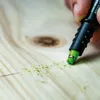 A hand is holding a black and green marker over a wooden surface, with green shavings scattered around the tip of the marker. Anteprima dell’immagine prodotto