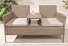 A wicker garden bench with a built-in table and a glass of water on it, set on a wooden deck with plants in the background. Produktbild-Vorschau