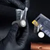 Hands wearing black gloves are holding a cylindrical object, possibly a battery, and a clear plastic bag. Anteprima dell’immagine prodotto