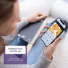 A person is holding a smartphone displaying a baby monitor app, with a baby visible on the screen. The person is sitting with a book. Produktbild-Vorschau