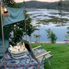 A camping scene by a lake with a folding chair and a tub of drinks. Produktbild-Vorschau