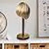 A desk lamp with a gold shade and a dark stand sits on a white shelf above wooden drawers. A circular object is partially visible to the left, and a framed picture is on the wall to the right. Produktbild-Vorschau