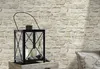 A black lantern with candles and stones sits on a wooden surface in front of a stone-patterned wallpaper. Produktbild-Vorschau