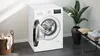 A white Siemens washing machine with its door open, placed in a laundry room setting with a basket of clothes and a potted plant. Produktbild-Vorschau