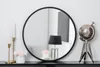 A round black-framed mirror is displayed on a black surface against a white wall. Decorative items like candlesticks, a vase of flowers, and a globe are placed around it. Produktbild-Vorschau