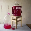 Two large glass carboys, one filled with red liquid and connected by a tube to the other, which is partially filled with red liquid. A small red airlock is in the foreground. The carboys are set up for fermentation or siphoning. Produktbild-Vorschau