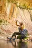 A woman is drinking from a water filter straw while sitting by a body of water with a backpack next to her, in a rocky, desert-like environment. Podgląd zdjęcia produktu