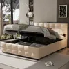 A bedroom scene featuring a beige upholstered bed with its storage compartment open, revealing stored items. A floor lamp is positioned behind the bed, and a window with blinds is visible to the left. Produktbild-Vorschau