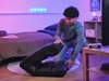 A young person is assembling a black floor chair in a bedroom with purple and pink lighting. Náhľad obrázku produktu