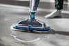 A person is using a cordless floor mop on a tiled floor, with water and cleaning solution being dispensed. Produktbild-Vorschau