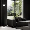A modern living room with a large window overlooking a forest and mountains. A dark gray sofa with pillows is in the foreground, and a tall, slender floor lamp stands to the left. Produktbild-Vorschau