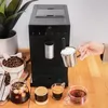 A coffee machine is shown on a wooden counter, with coffee beans and cups of coffee around it. A hand is pouring frothed milk into a cup. Produktbild-Vorschau
