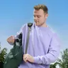 A young man is putting a phone into a green backpack while wearing a purple sweatshirt. Produktbild-Vorschau