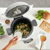 A person is adding liquid to a multicooker with food inside, surrounded by ingredients. Produktbild-Vorschau