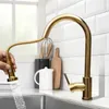 A gold kitchen faucet with a pull-down sprayer is shown in use over a stainless steel sink. The background features white subway tiles and kitchen accessories. Podgląd zdjęcia produktu