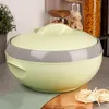 A pale green and gray insulated food container sits on a wooden cutting board, with a green cloth and uncooked pasta in the foreground. Jars of dry goods and pasta are visible in the background. Produktbild-Vorschau