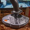 A close-up of a ThrustMaster SimTask FarmStick X joystick on a wooden surface, with a blurred background showing a screen with a game interface. Anteprima dell’immagine prodotto