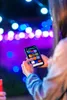 A person is holding a smartphone with a colorful interface, likely controlling lights in the background. Náhled obrázku produktu