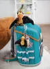 A teal backpack with orange trim, decorated with plush dinosaur toys hanging from a zipper. Produktbild-Vorschau