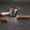 A coffee portafilter filled with coffee grounds and a coffee distribution tool with wooden handles and metal needles are placed on a dark surface. Produktbild-Vorschau