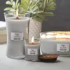 Three lit WoodWick candles of varying sizes are arranged on a surface, with a plant in the background. Produktbild-Vorschau