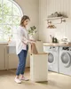A woman is putting laundry into a tall, beige hamper with a wooden lid in a laundry room. Náhled obrázku produktu