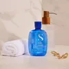 A blue bottle of shampoo and a rolled-up white towel are placed on a marble surface, with a dispenser in the background and gold jewelry in the foreground. Anteprima dell’immagine prodotto