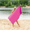 A woman holding a pink and blue towel on a sandy beach by a lake. Produktbild-Vorschau