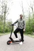 A man stands on an electric scooter on a paved path in a wooded area. Náhled obrázku produktu