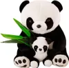 Two plush panda toys, one large and one small, are sitting together. The larger panda is behind the smaller one and is holding a few green bamboo leaves. Produktbild-Vorschau